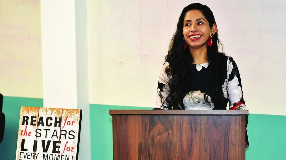 A woman stands at a wooden podium, smiling, with a motivational sign beside her that reads, "Reach for the stars, live for every moment.