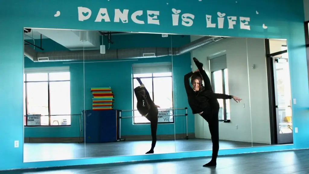 Una bailarina vestida de negro realiza un estiramiento de piernas altas en un estudio de danza con "Dance Is Life" escrito en la pared encima de un gran espejo.
