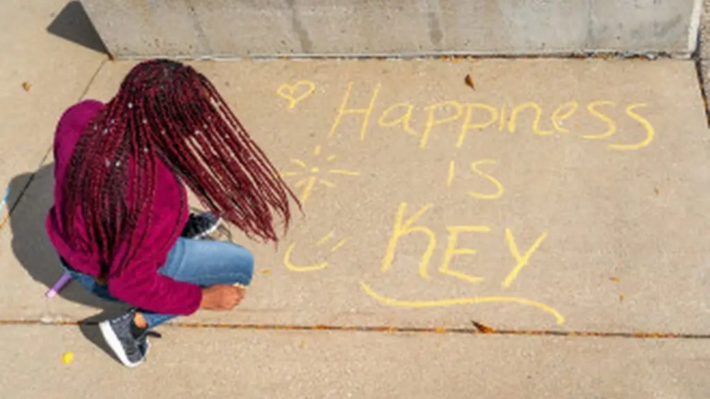 A person with braided hair writes "Happiness is Key" in yellow chalk on a sidewalk next to a concrete wall.