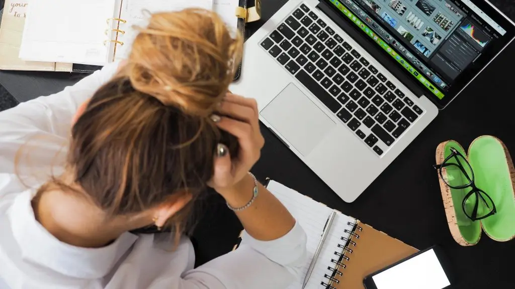 Person with a messy bun sits at a desk with an open laptop, notebooks, glasses case, and phone, holding their head in one hand and appearing stressed or frustrated.