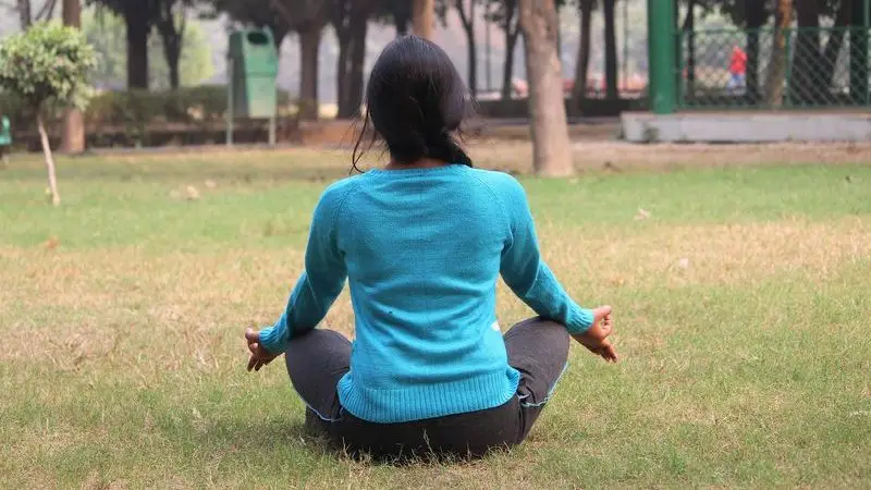 Una persona se sienta con las piernas cruzadas sobre la hierba de un parque, mirando hacia otro lado, con las manos apoyadas en las rodillas en postura de meditación.