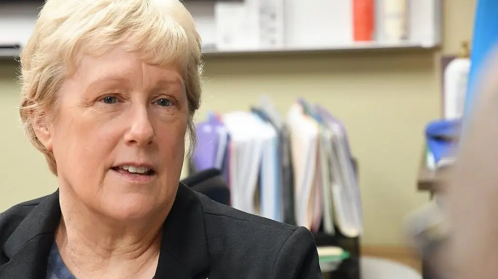 A person with short blonde hair and a black blazer is sitting indoors with shelves and organized files in the background.