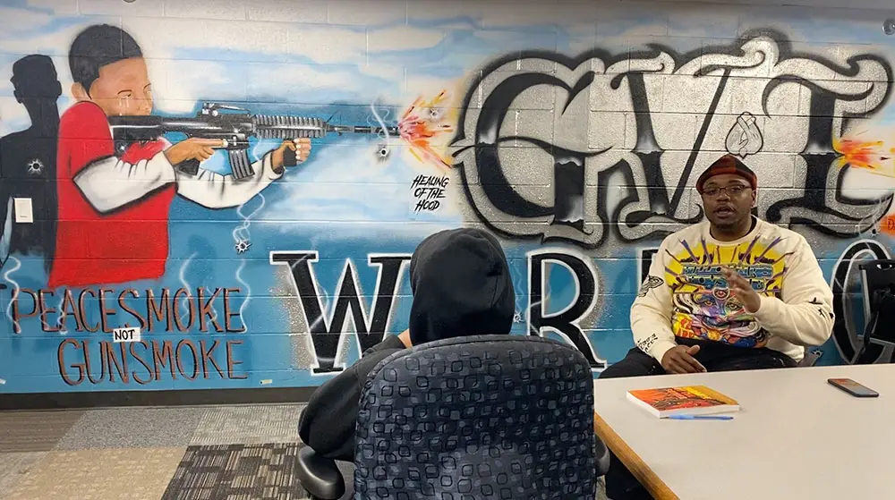 Dos personas conversan en una mesa frente a un mural que muestra a una persona con una pistola, texto en graffiti y la frase "Peacesmoke not Gunsmoke".