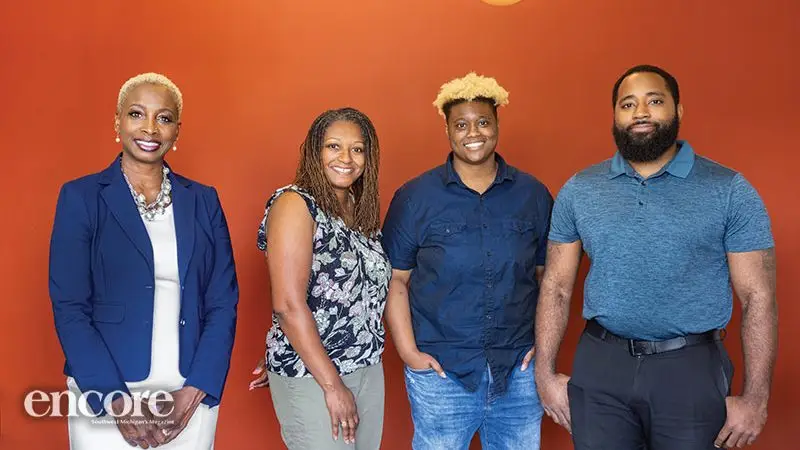 Four adults stand side by side in front of an orange wall, facing the camera. The "encore" logo appears in the lower left corner.