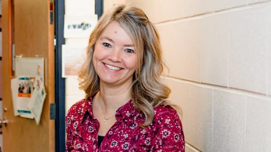 A woman with long blonde hair wearing a red floral shirt smiles while standing in a hallway with white brick walls.