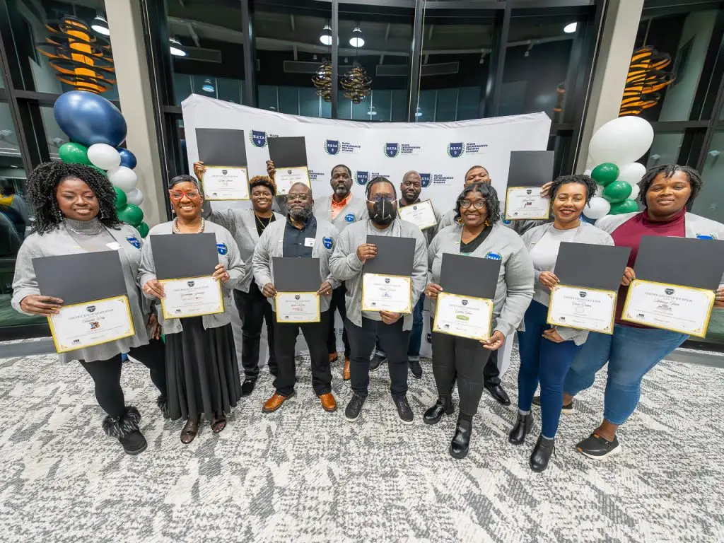 A group of thirteen adults stands indoors holding certificates and smiling at the camera, with balloons and a branded backdrop behind them.