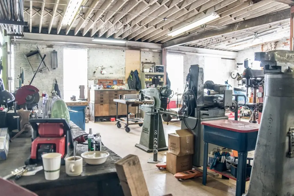 A workshop with various industrial machines, tools, workbenches, scattered supplies, and fluorescent lighting in a spacious, unfinished room.