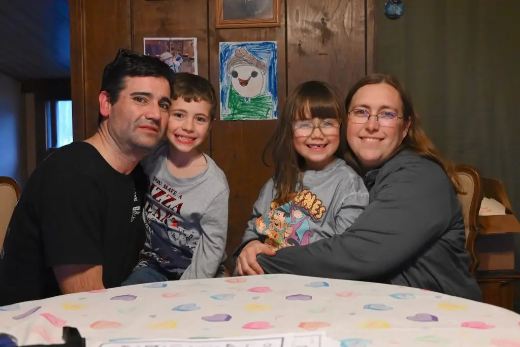 A man, boy, girl, and woman sit closely together at a table with a heart-patterned tablecloth, smiling for the camera. Children's drawings are visible on the wall behind them.