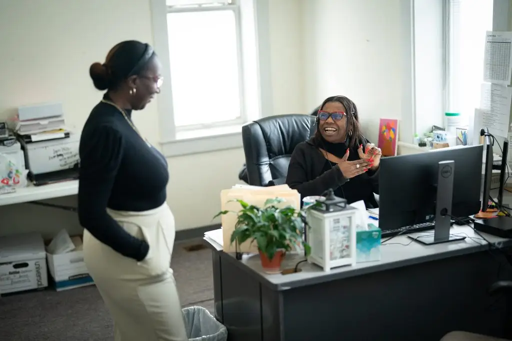 Two women interact in an office; one is sitting at a desk with a computer, smiling and gesturing, while the other stands nearby.