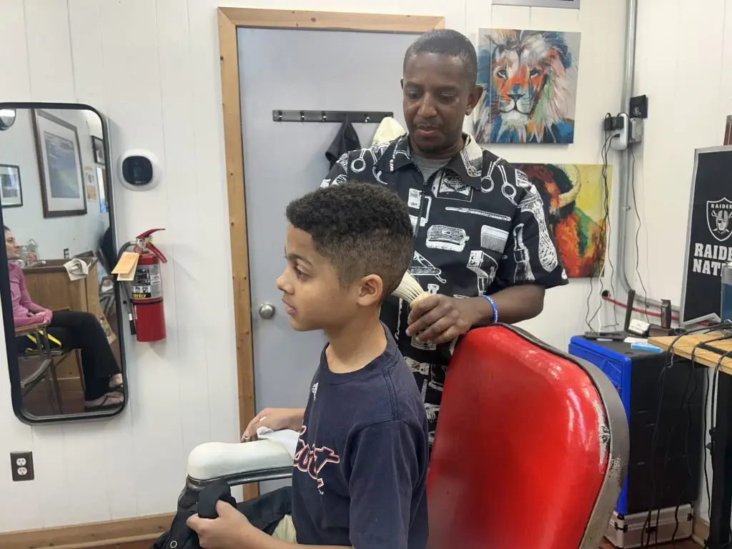 A young boy sits in a barber chair while a barber prepares to cut his hair in a barbershop decorated with art and sports memorabilia.