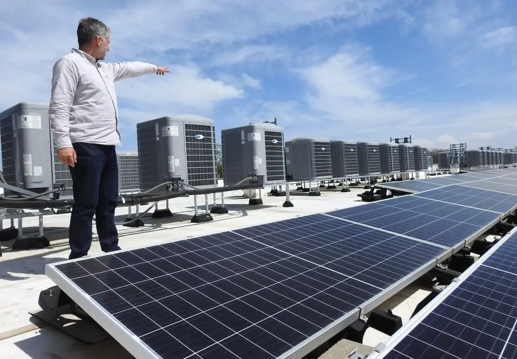 A man stands on a rooftop near solar panels and air conditioning units, pointing toward the row of HVAC systems under a partly cloudy sky.