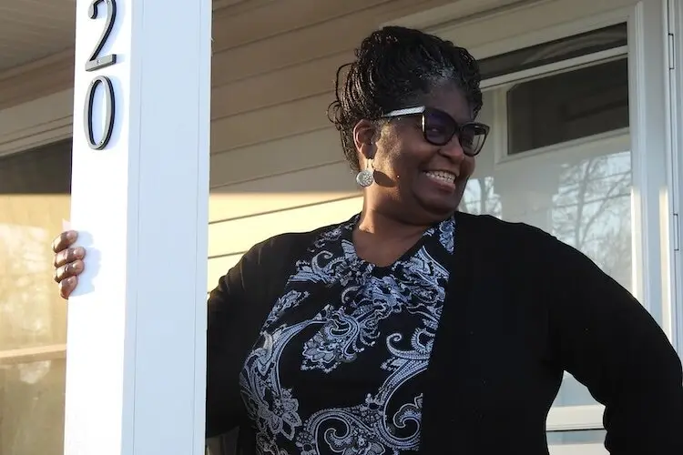 A woman wearing glasses and a patterned top stands outside a house, smiling and resting her hand on a post with the number 20.
