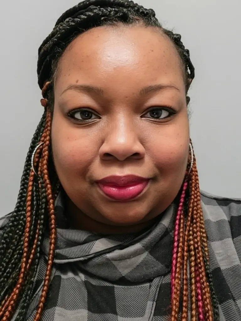 A woman with braided hair and hoop earrings, wearing a gray plaid top, looks directly at the camera against a plain light gray background.