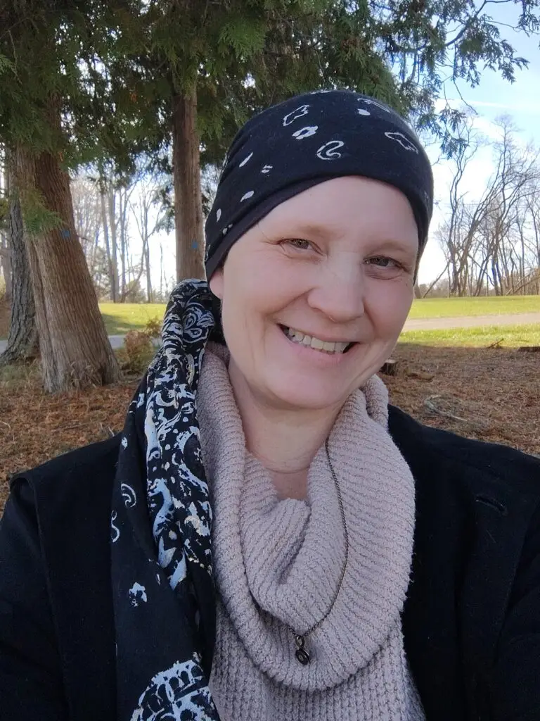 A smiling person wearing a patterned headscarf, knit scarf, and dark coat poses outdoors near large trees and grass.