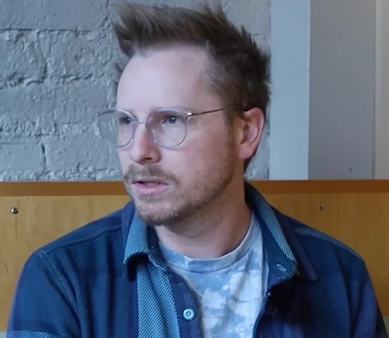 A person with glasses and short, spiked hair is sitting indoors against a light-colored brick wall, wearing a blue plaid shirt over a tie-dye t-shirt.