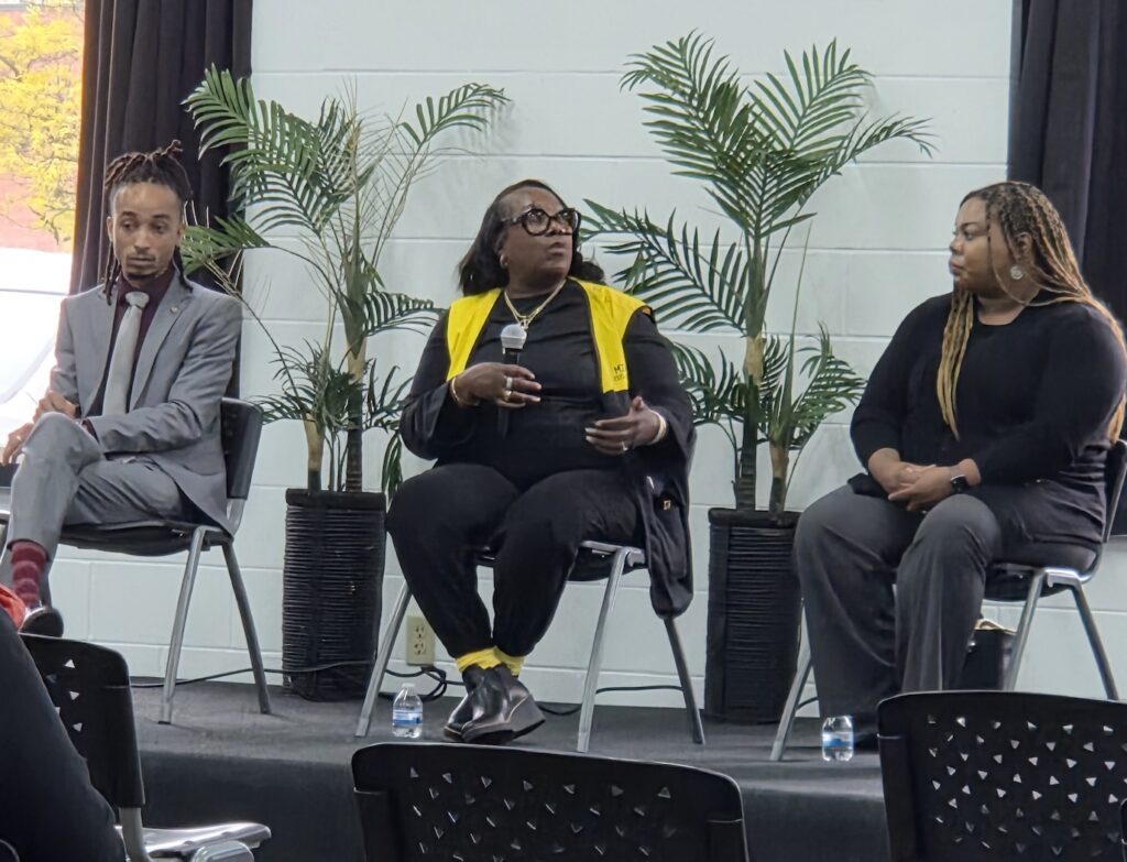 Three people sit on a stage in front of potted plants, engaging in a panel discussion. Audience members are partially visible in the foreground.