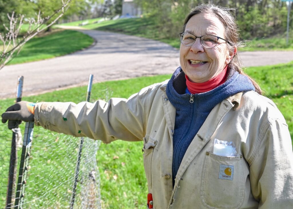 A person wearing glasses and work gloves stands by a chain-link fence outdoors, with a winding road and trees in the background on a sunny day.