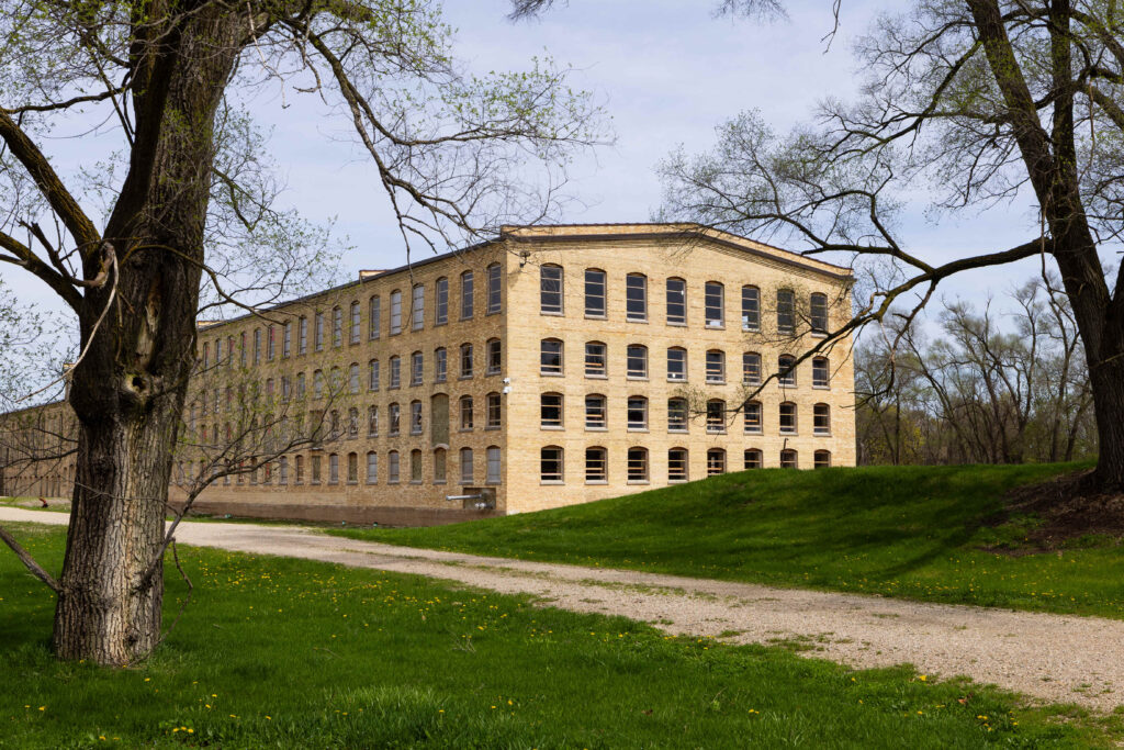 A large, rectangular, four-story brick building with many windows stands on a grassy area, bordered by trees and a gravel path.