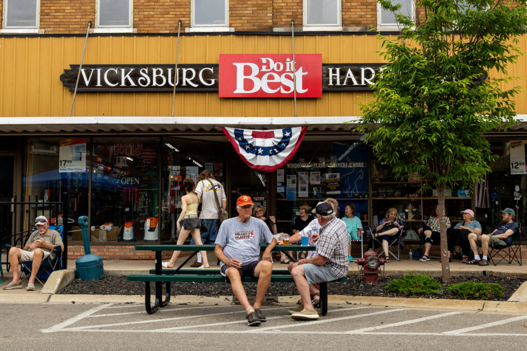Several people sit on benches outside a hardware store with a "Vicksburg Do it Best Hardware" sign and patriotic banner, while others gather near the entrance on a cloudy day.
