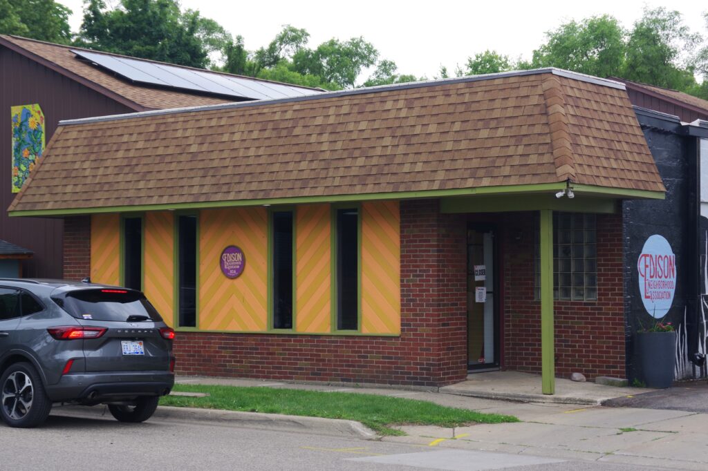 A small brick building with a brown roof and yellow-orange patterned panels, displaying signs for "Fusion" on its window and wall; a parked SUV is nearby.