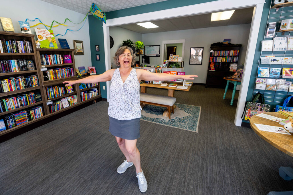 A woman stands in a bookstore with arms outstretched, smiling. Bookshelves, tables, and greeting card displays are visible in the background.