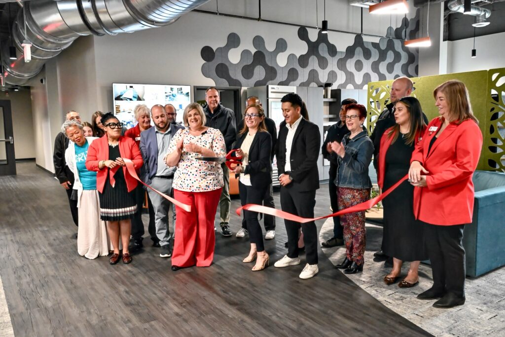 A group of people stands in an office space as a woman in the center cuts a red ribbon for a ribbon-cutting ceremony.