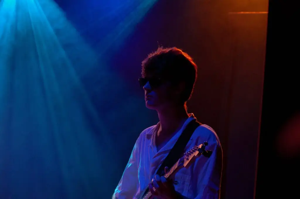 A person wearing sunglasses and a white shirt plays guitar on stage under blue and orange lights.