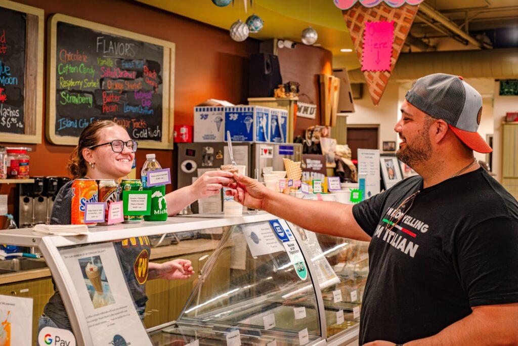 A man receives an ice cream sample from a smiling employee behind the counter at an ice cream shop, with a menu board and drinks visible in the background.