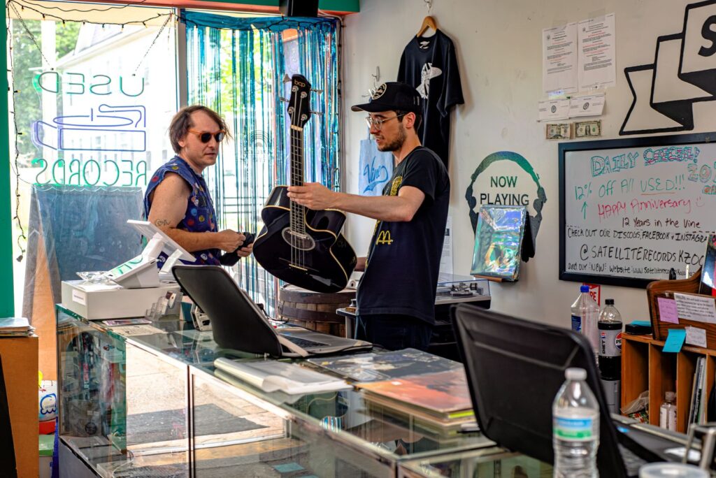 Two men stand behind a record store counter; one hands an acoustic guitar to the other. Records, a t-shirt, and sale signs are visible in the colorful, cluttered shop.