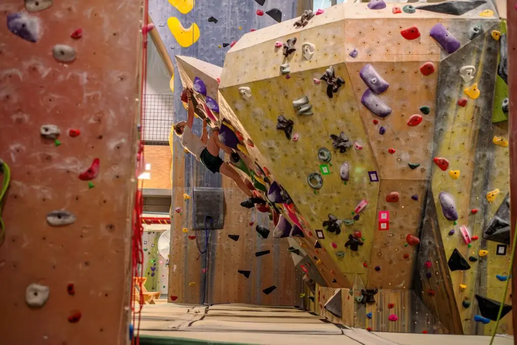 A person climbs an indoor bouldering wall with colorful holds and padded flooring visible in a rock climbing gym.