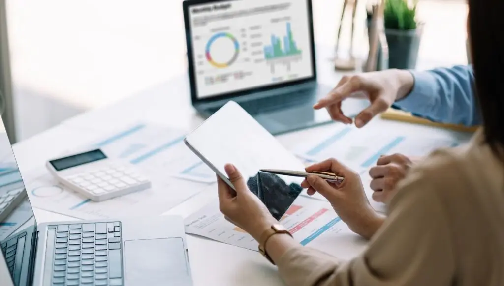 Two people review financial charts on a tablet and printed documents at a desk, with laptops and a calculator visible in the background.