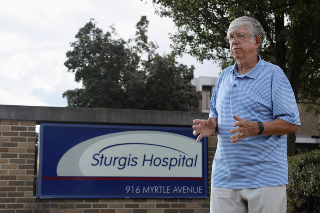 An older man stands in front of a Sturgis Hospital sign at 916 Myrtle Avenue, with trees and part of the hospital building visible in the background.