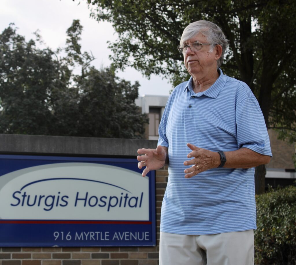 An older man stands in front of a Sturgis Hospital sign at 916 Myrtle Avenue, with trees and part of the hospital building visible in the background.