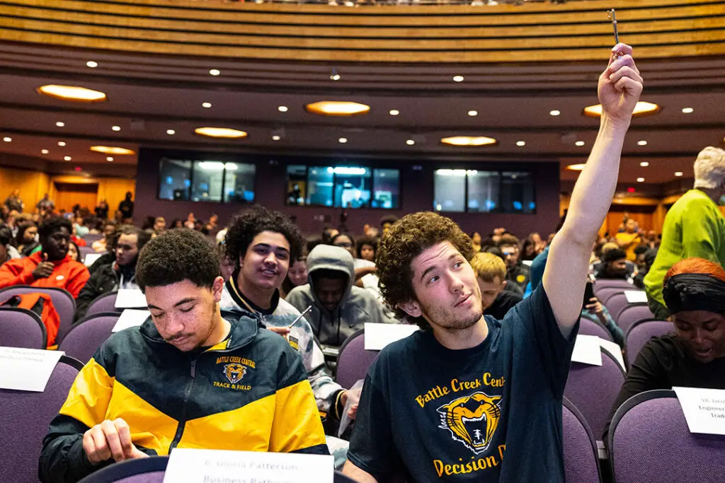 A young man in a blue t-shirt raises his hand in a crowded auditorium, surrounded by other seated students.